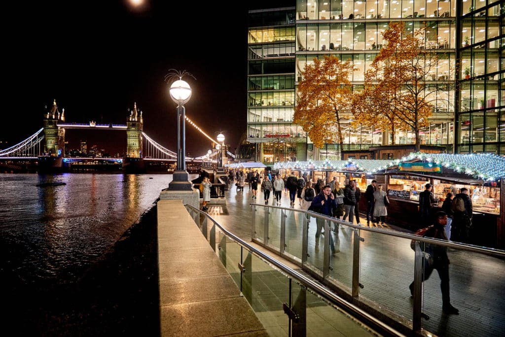 a shot of christmas by the river, which starts in november, with people walking along the river next to twinkling light fronted stalls