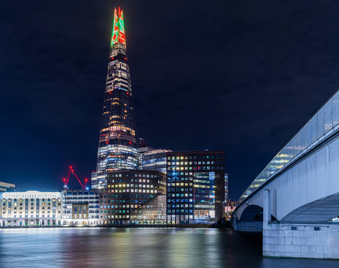A distant shot of the shard at night, amongst other London buildings, with a christmas light display taking over the top portion of the building