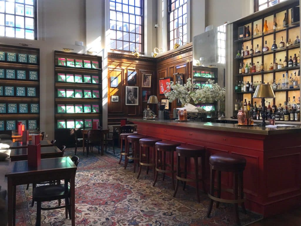 the swans bar at maison assouline, a wooden bar with stools lined up against it nestled in between the store's boolshelves